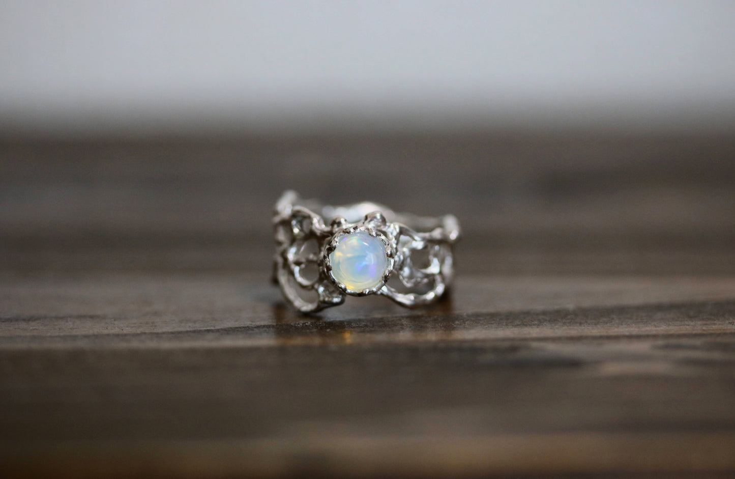 A silver ring with an irregular, organic-shaped setting and a central iridescent white opal gemstone, resting on a dark wooden surface.