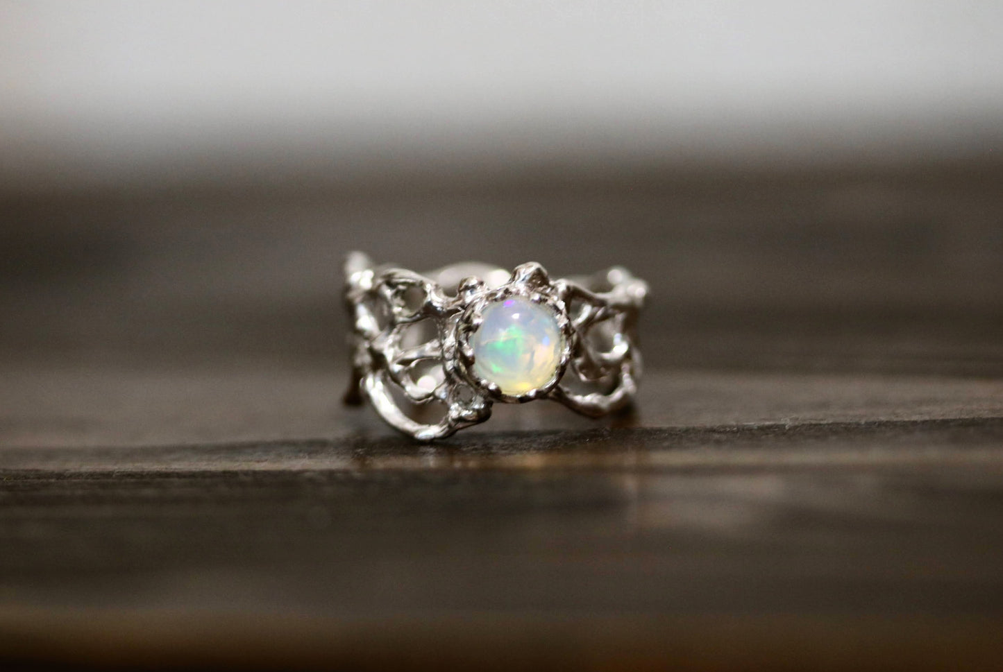 A silver ring with an ornate, textured band and a central round iridescent opal gemstone, resting on a dark wooden surface.