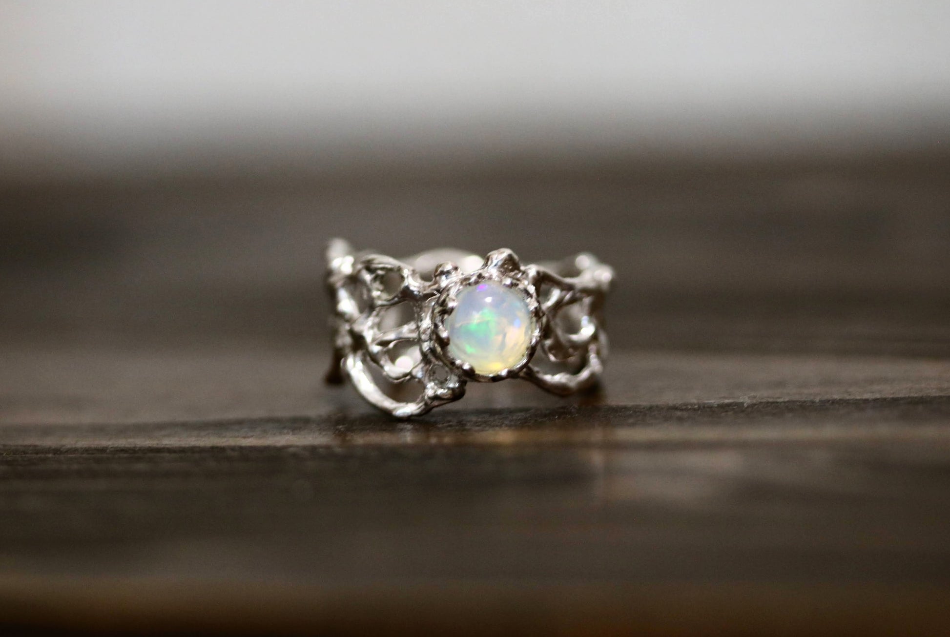 A silver ring with an ornate, textured band and a central round iridescent opal gemstone, resting on a dark wooden surface.