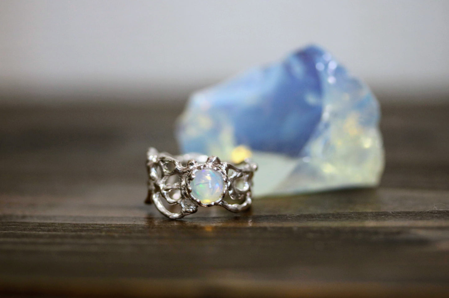 A silver-toned ring with a central round iridescent opal gemstone, displayed on a dark wooden surface with a blurred blue iridescent stone in the background.