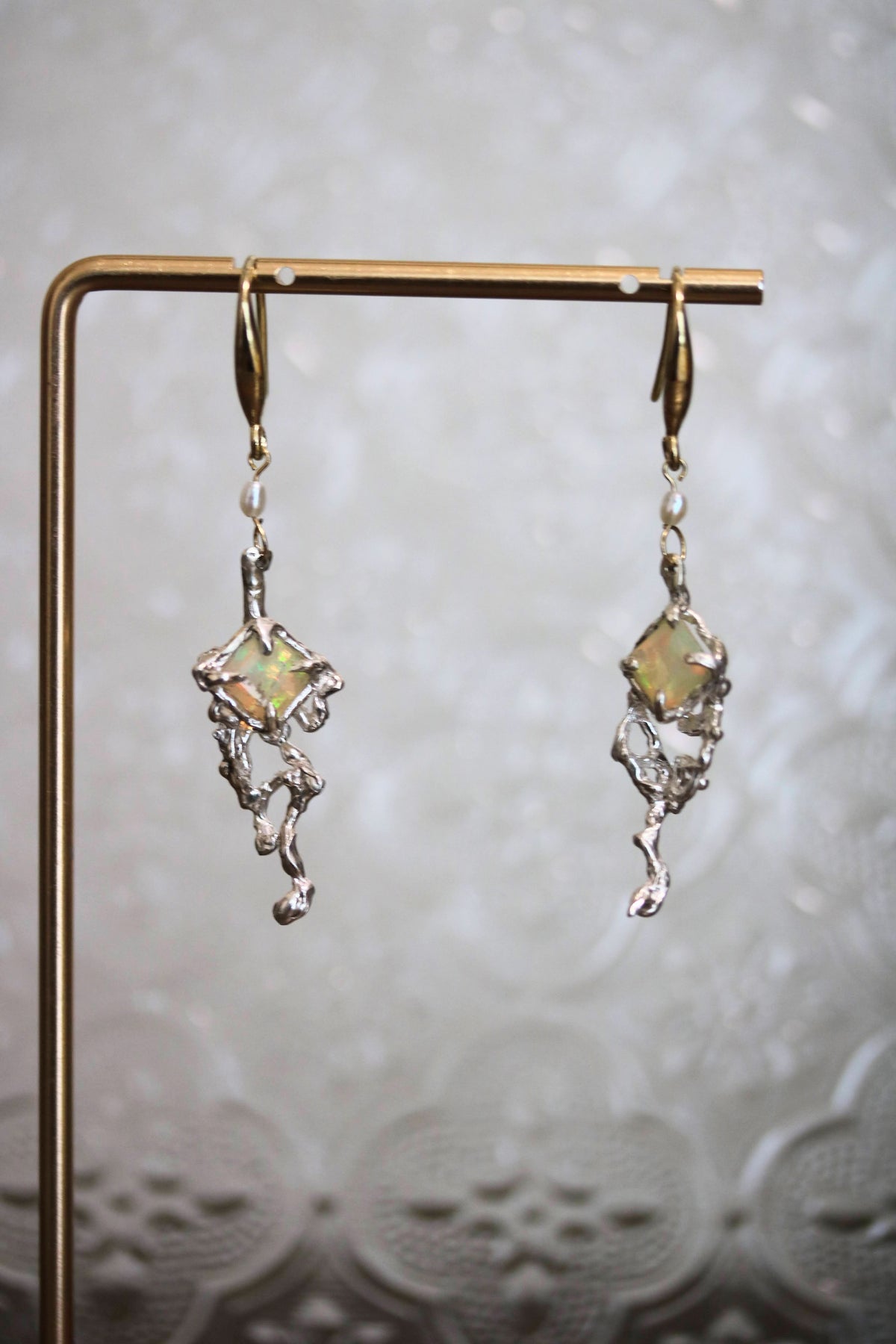 A pair of dangling earrings displayed on a gold-tone stand against a soft, textured light gray background. The earrings feature silver-toned organic-shaped metalwork with a central iridescent, opalescent square stone.