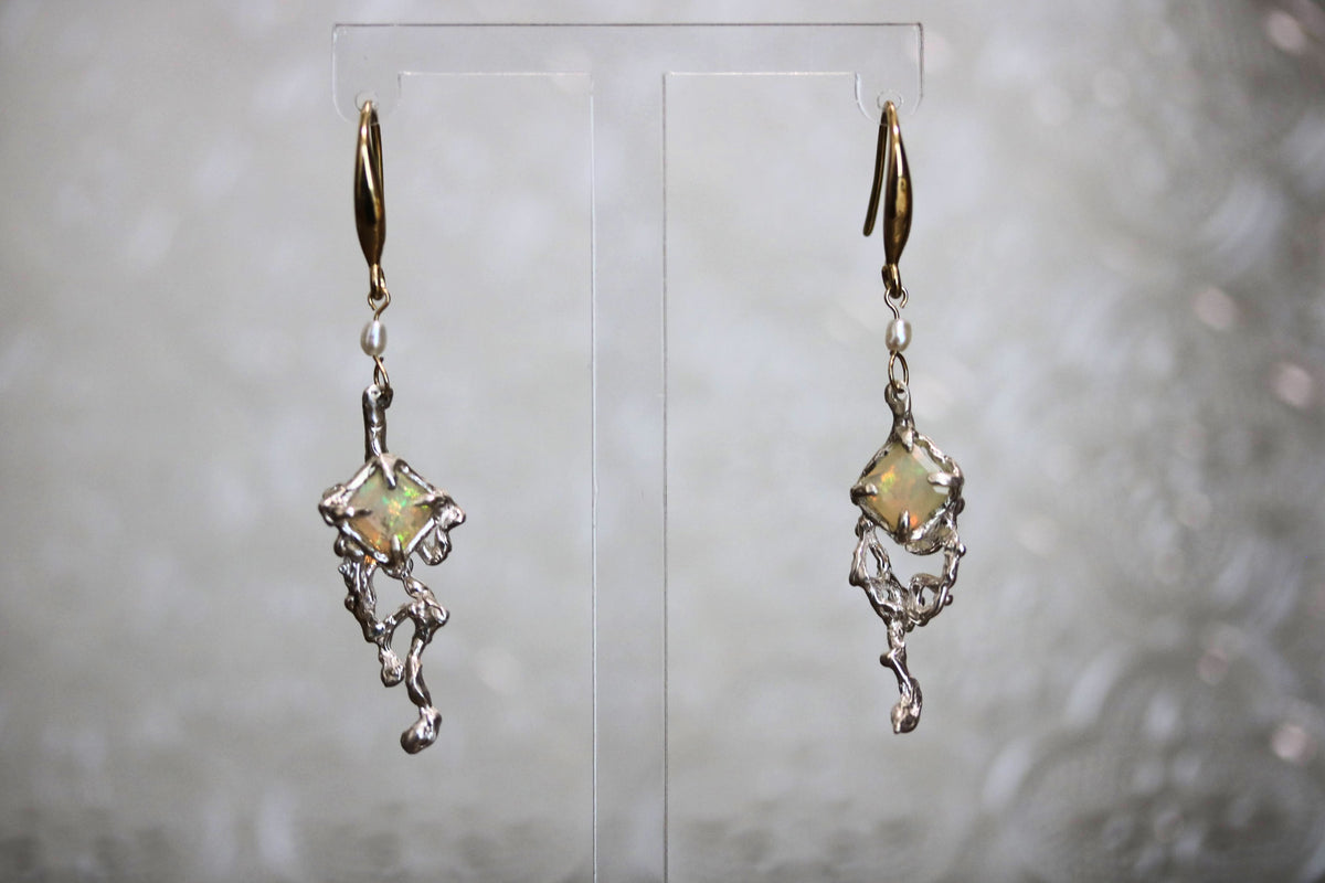 A pair of dangling earrings displayed on a clear acrylic stand against a soft, out-of-focus light gray background. Each earring features a small iridescent, star-like gemstone with a green and gold sheen, suspended from an organic, silver-toned, branch-like metal frame with gold-toned hooks at the top.