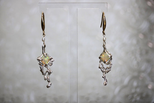 A pair of dangling earrings displayed on a clear acrylic stand against a soft, out-of-focus light gray background. Each earring features a small iridescent, star-like gemstone with a green and gold sheen, suspended from an organic, silver-toned, branch-like metal frame with gold-toned hooks at the top.