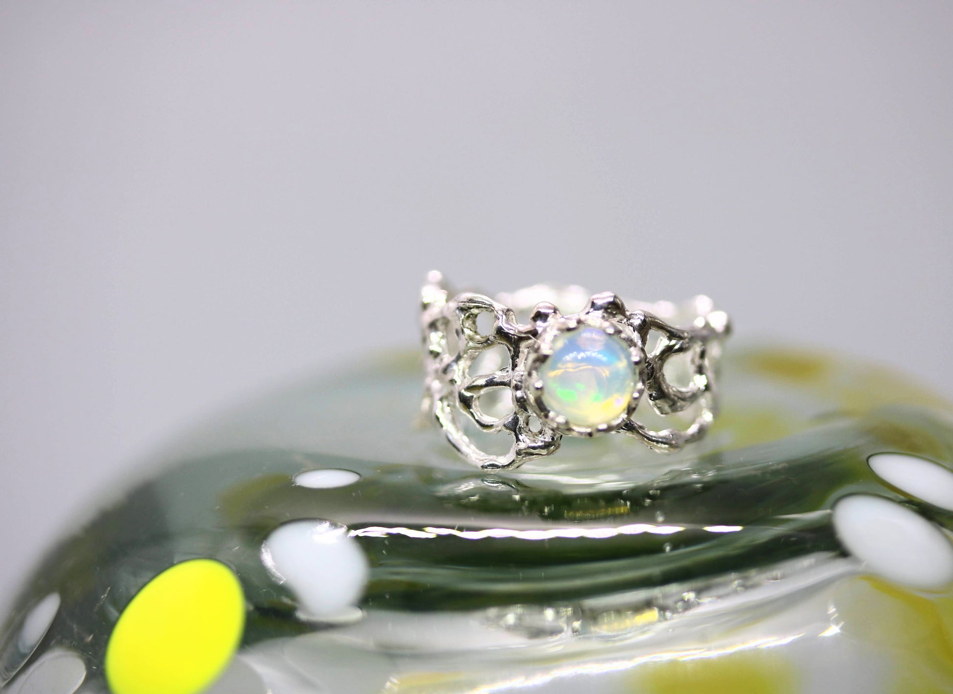 A silver ring with an oval iridescent opal center stone, displayed on a reflective green glass surface against a light gray background.