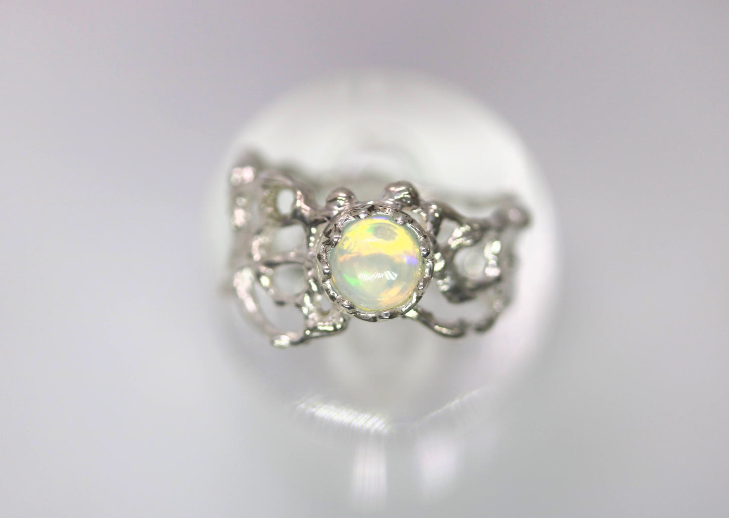 A silver ring featuring a round iridescent opal gemstone in a decorative filigree setting, displayed on a reflective white surface.