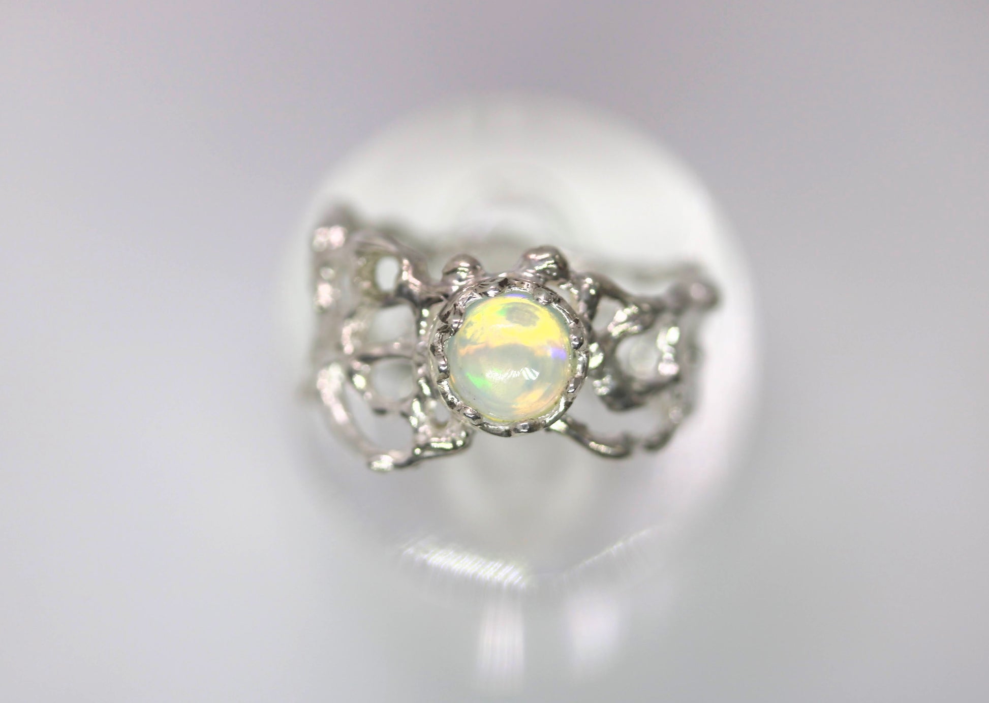 A silver ring featuring a round iridescent opal gemstone in a decorative filigree setting, displayed on a reflective white surface.