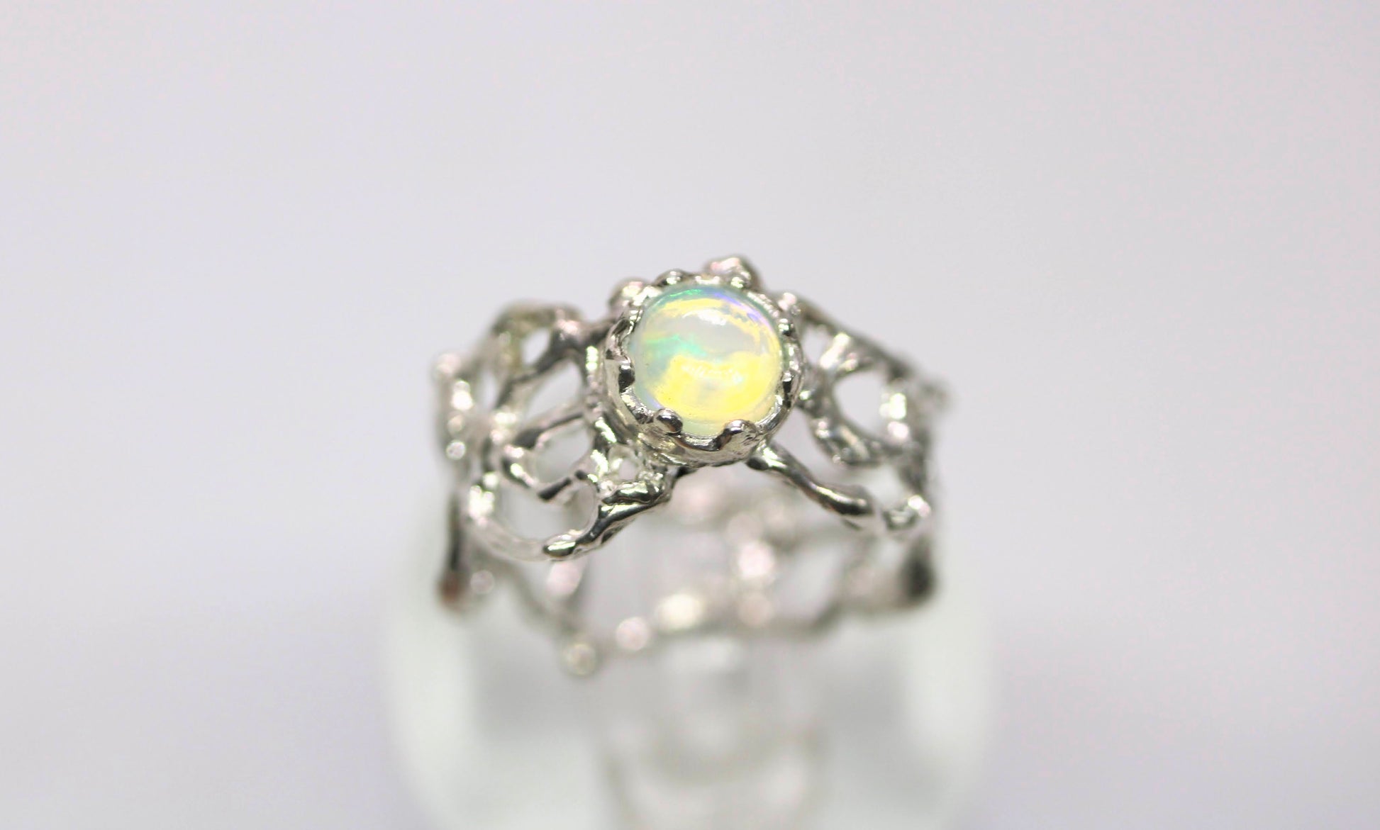 A silver ring featuring a round iridescent opal gemstone in a decorative filigree setting, displayed on a soft white surface against a light gray background.