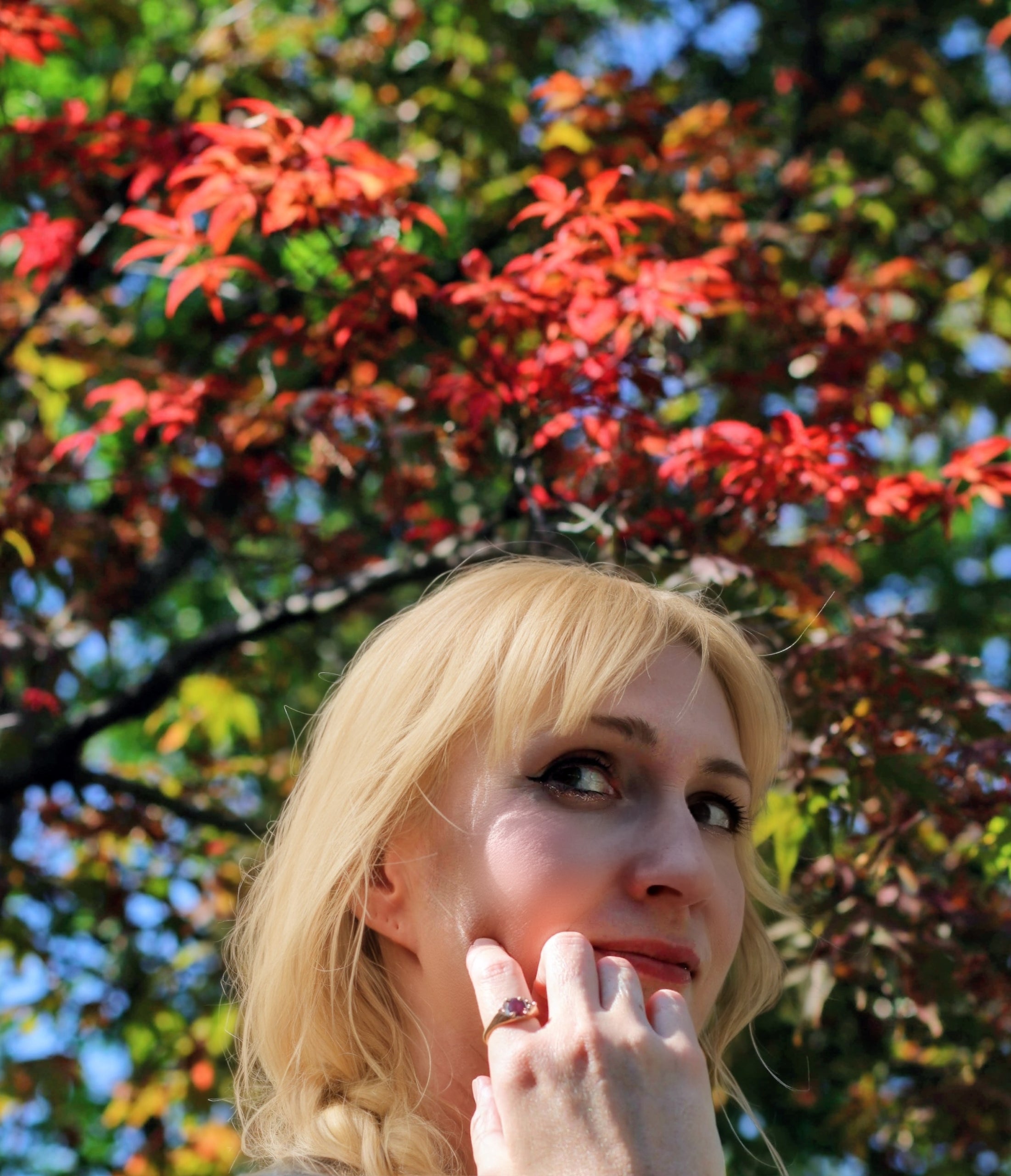 Woman with blonde hair and a ring, standing in front of a tree with red and green leaves.